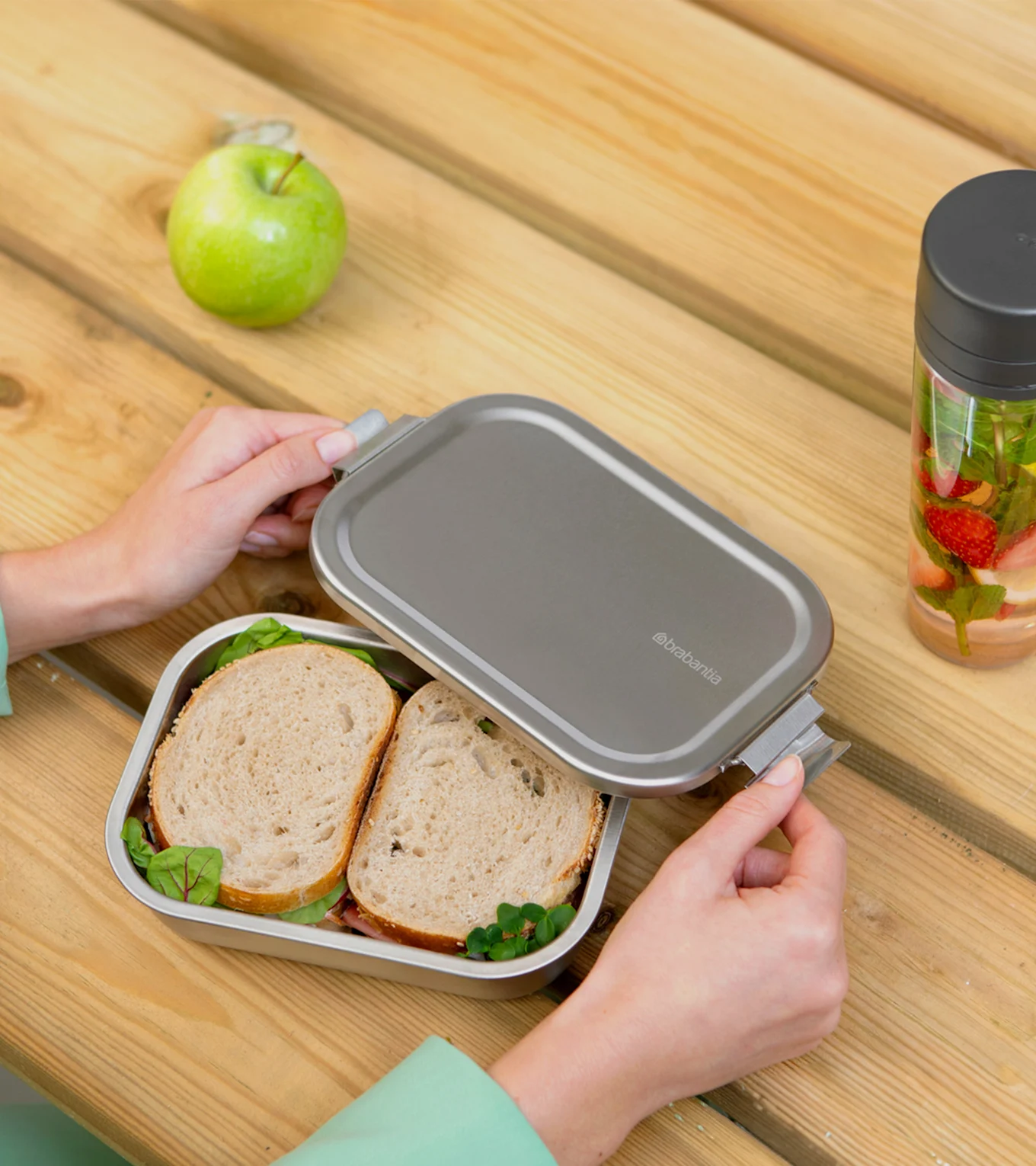 Hænder åbner en sølvfarvet madkasse med sandwich, et æble og en frugtvandflaske på et træbord.