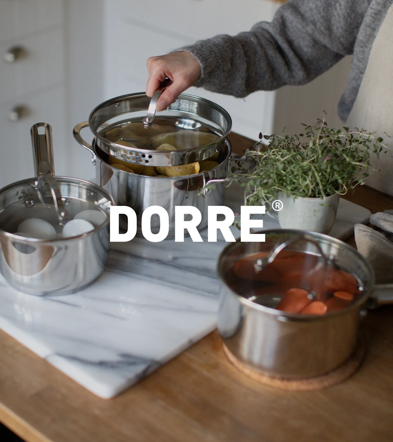 Hånd løfter låg fra rustfri stål damppotte med kartofler, ved siden af potter med æg og gulerødder. DORRE logo.